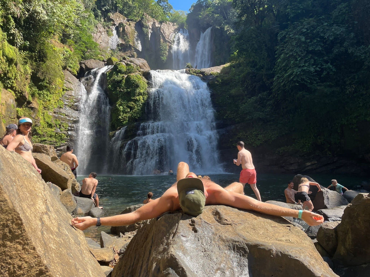 A person relaxes on a large rock in the foreground of a scenic waterfall in a tropical jungle setting at a Costa Rica Retreat with other people swimming and sitting around the pool below.