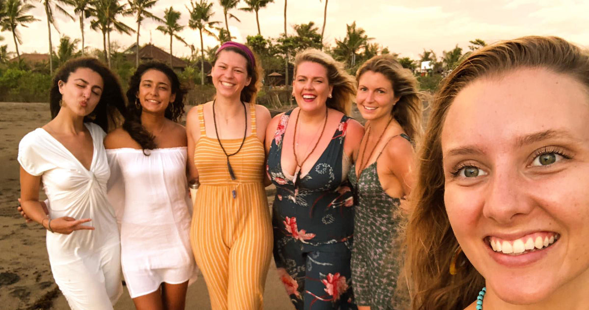 Five women posing together on a beach at sunset, wearing various colorful dresses and swimwear. The photo is a casual selfie with palm trees and sand in the background.