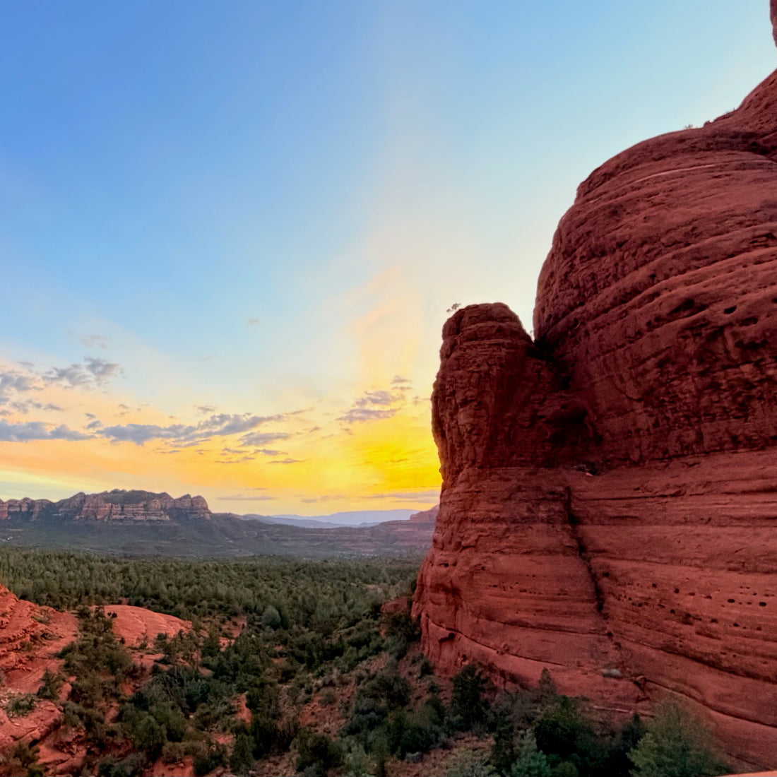 A scenic landscape photograph of red rock desert formations framing a valley at sunset, with a blue sky, warm yellow glow near the horizon, and green vegetation in the distance.