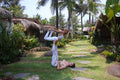 Two people practicing yoga on a tropical resort lawn with palm trees and thatched-roof bungalows in the background.