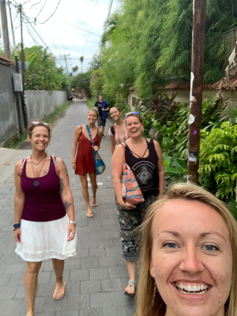 A group of women smiling and walking down a paved outdoor street lined with green trees and plants. The photo is taken in a casual selfie style, with one woman in the foreground and several others behind her wearing casual summer clothing.