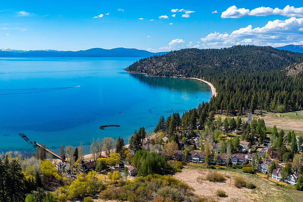 Aerial view of Lake Tahoe overlooking a Tahoe Retreat with turquoise water, a sandy bay, forested hills, and scattered houses under a blue sky with white clouds.